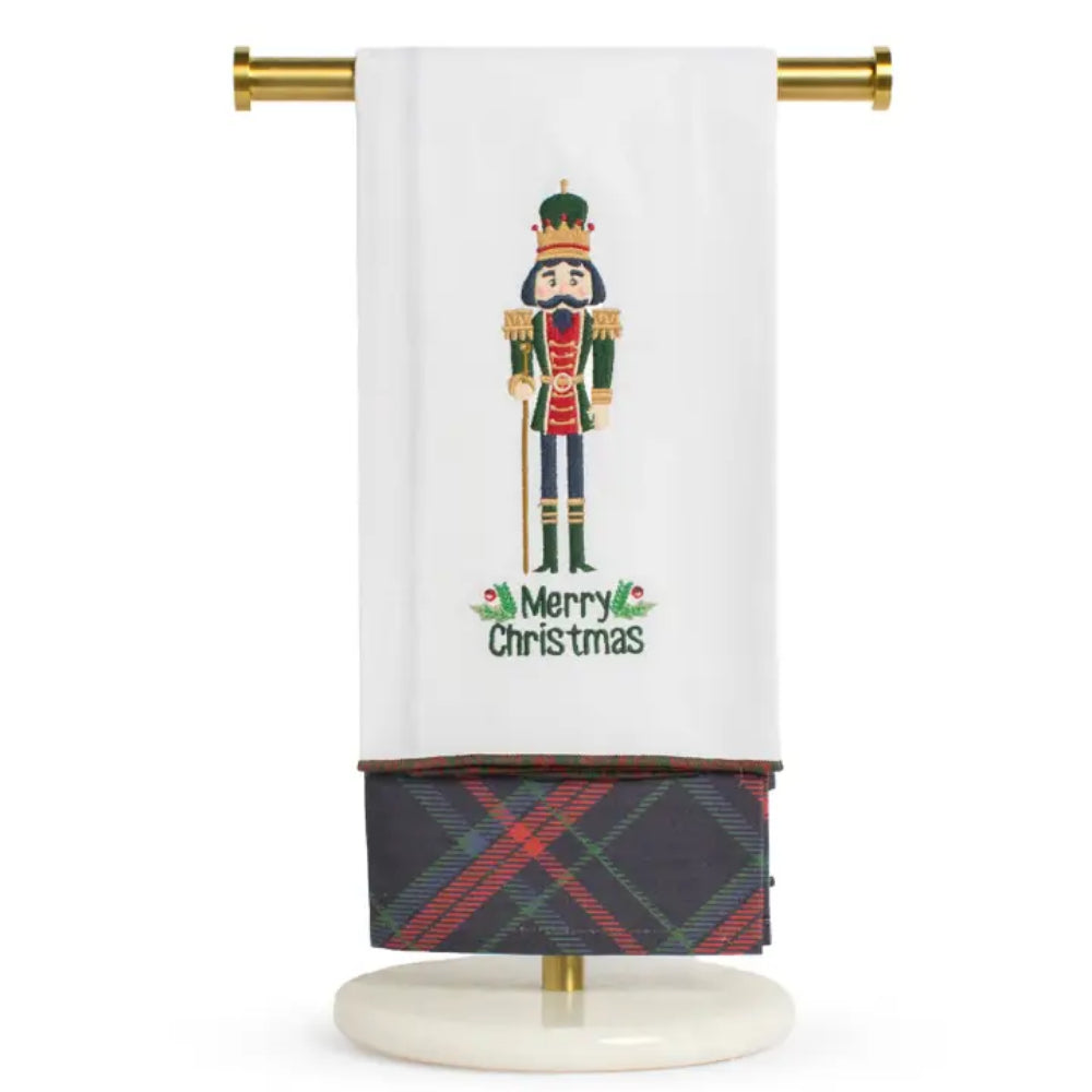 A white Christmas-themed towel hanging from a gold rod on a stand. The towel features a colorful nutcracker illustration and the words 'Merry Christmas' in green, with a folded section at the bottom showing a red, green, and black tartan plaid pattern.