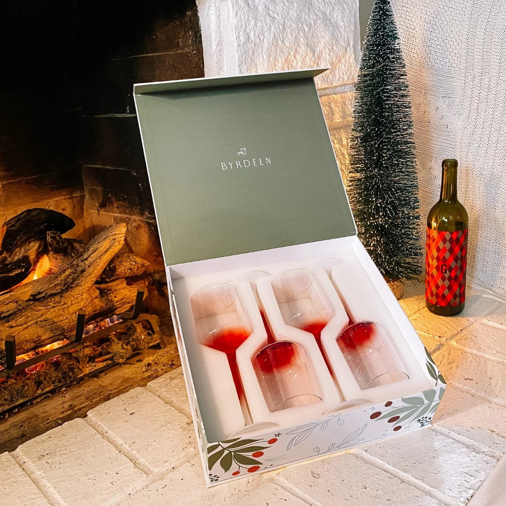A boxed set of four Byrdien wine glasses with deep red interiors, presented in an open gift box on a white brick surface beside a fireplace and a small decorated tree.