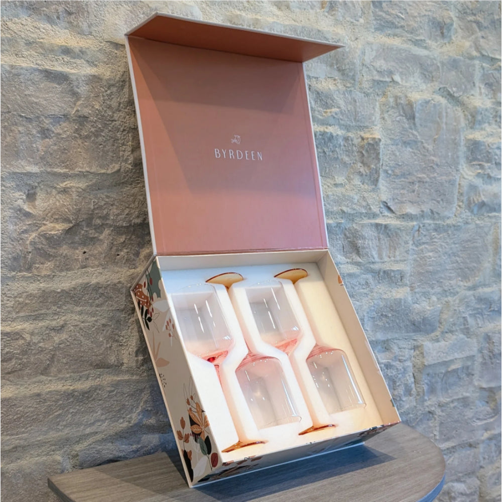 A boxed set of four Byrdeln clear stemmed glasses with pink liquid inside, displayed in an open pink presentation box against a gray stone wall.