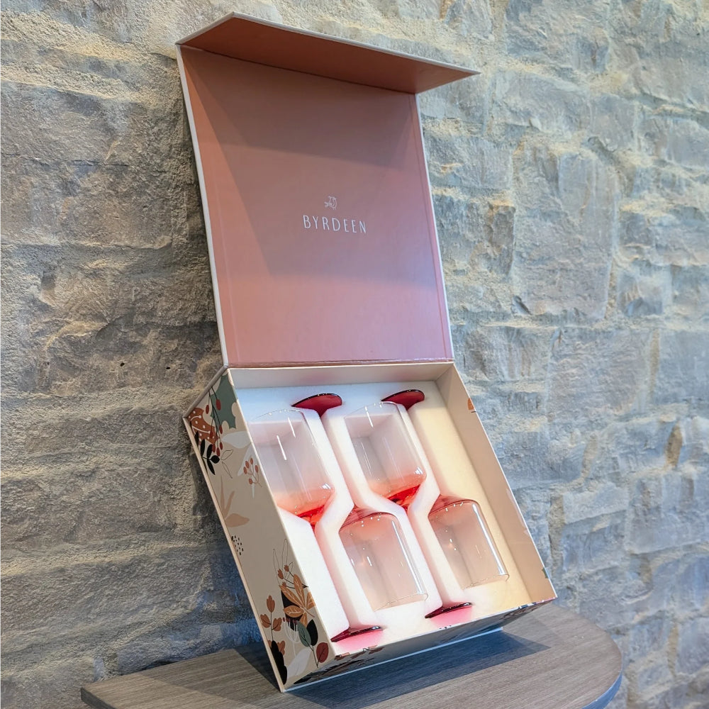 A boxed set of four Byrdeen stemmed wine glasses displayed in an open presentation box, set against a rustic stone wall background.