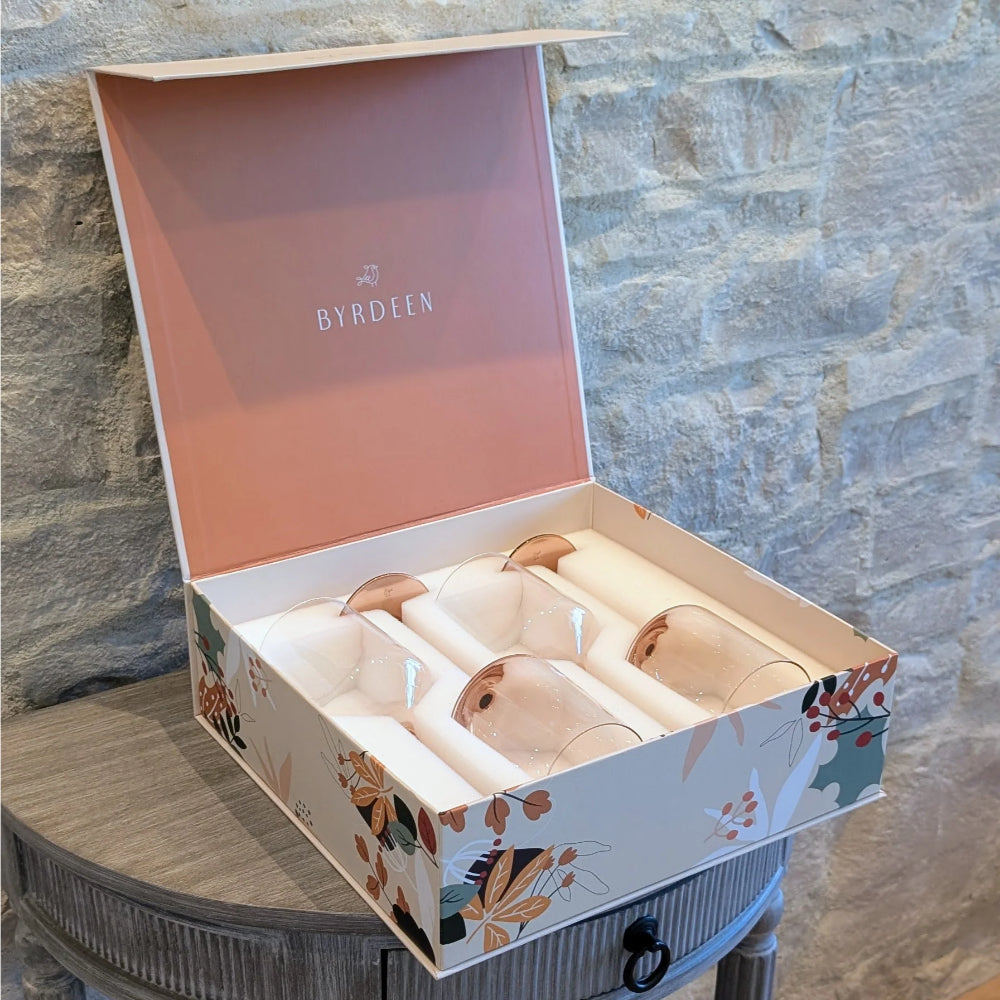 An open BYRDEEN gift box containing four clear glass tumblers with rose gold rims, displayed on a rustic gray table against a stone wall background.