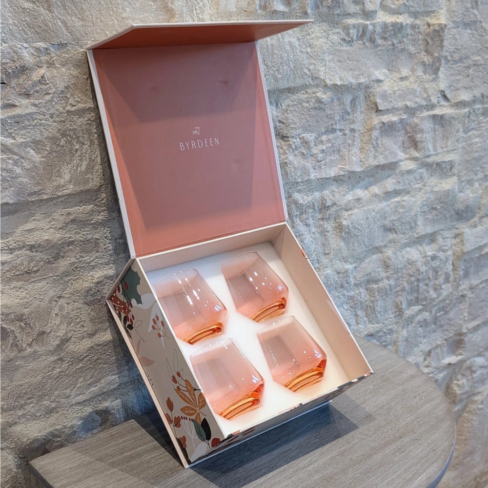 A boxed set of four pink-tinted glass tumblers presented in an open decorative gift box, photographed against a stone wall and tabletop.