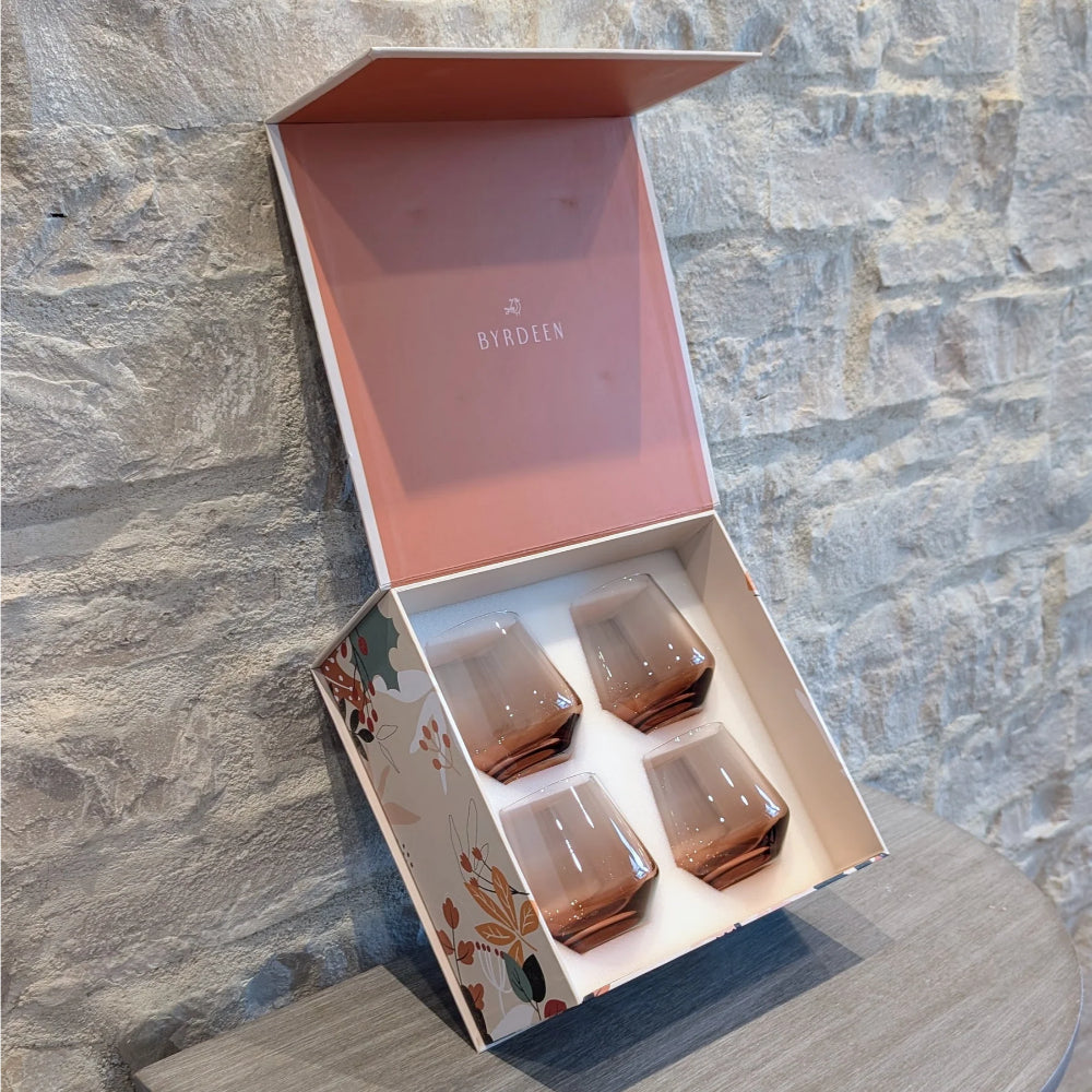 An open Birden gift box containing four amber-tinted lowball glasses, set against a gray stone wall background.