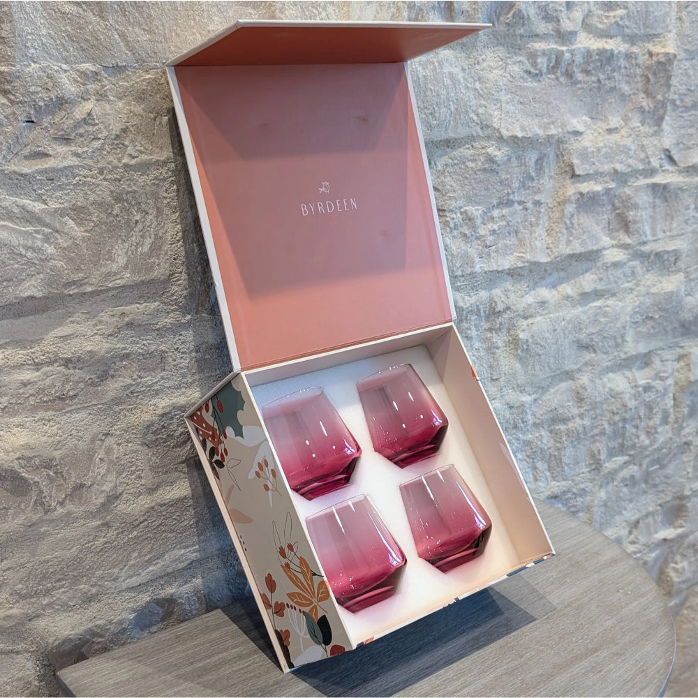 An open BYRDEEN gift box containing four pink gradient glass set against a rustic stone wall background.
