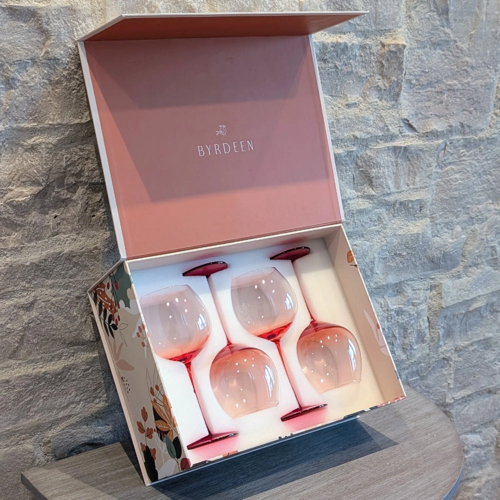 A boxed set of four Byrdeen stemmed wine glasses with pale pink bowls and deeper pink stems, presented in an open floral gift box against a stone wall.