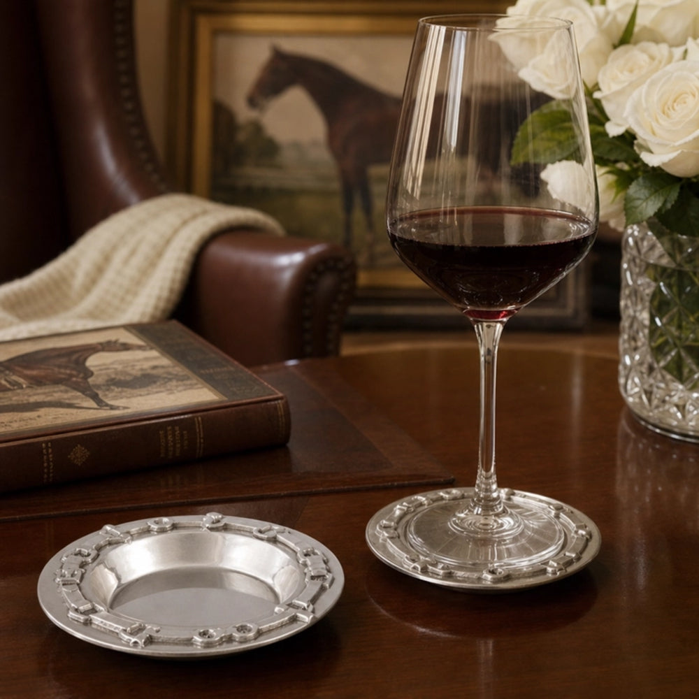 A silver-toned engraved wine glass coaster and matching small plate set on a dark wooden table, with a filled wine glass standing beside them in a styled indoor setting.
