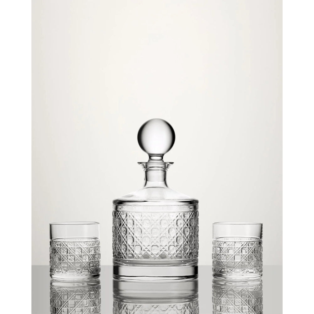 A clear glass decanter with a round stopper and two matching cut-glass tumblers, arranged on a reflective surface against a light neutral background.