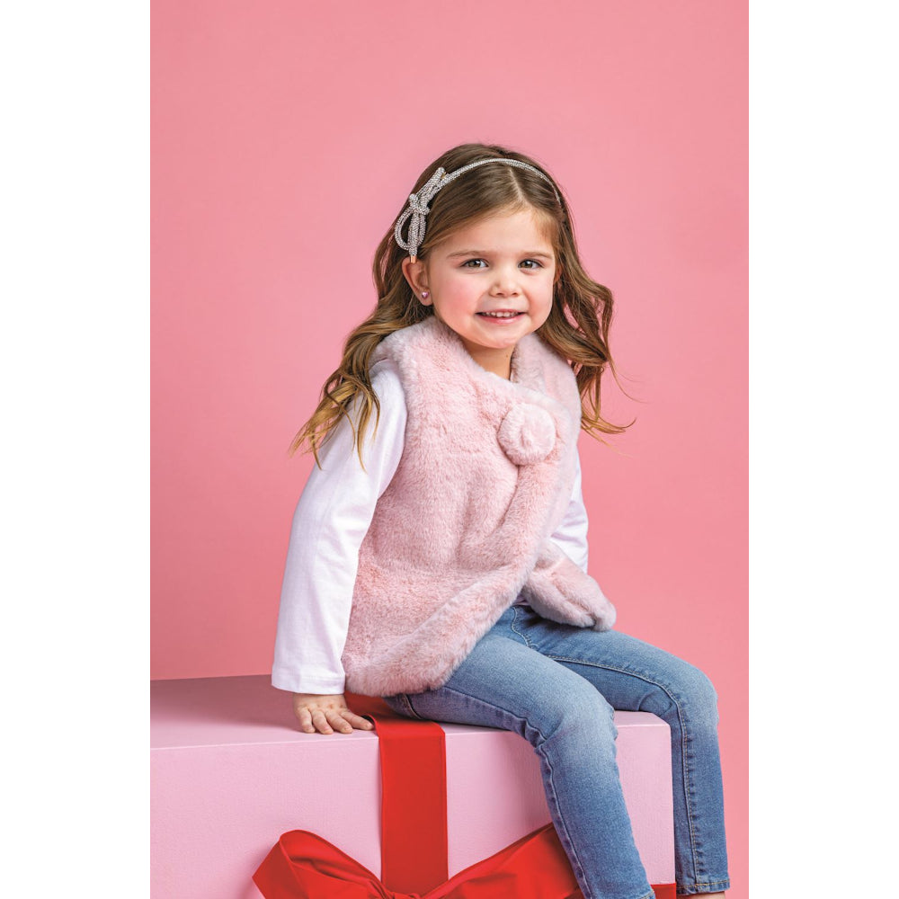 Young girl wearing a pink fur vest sitting on a pink box with a red bow against a pink background.