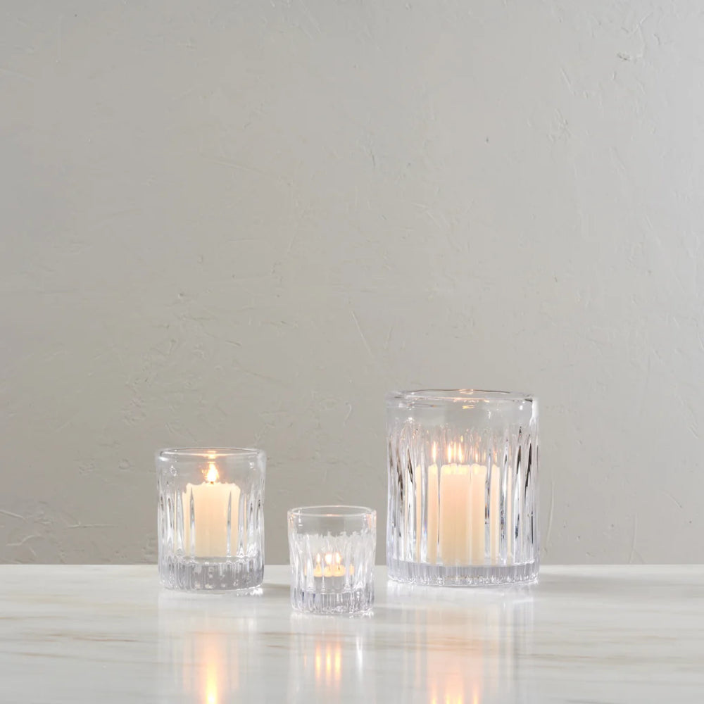 Three clear glass candle holders with vertical fluted detailing, each containing a lit cream-colored pillar candle, arranged on a light marble surface against a neutral textured wall.