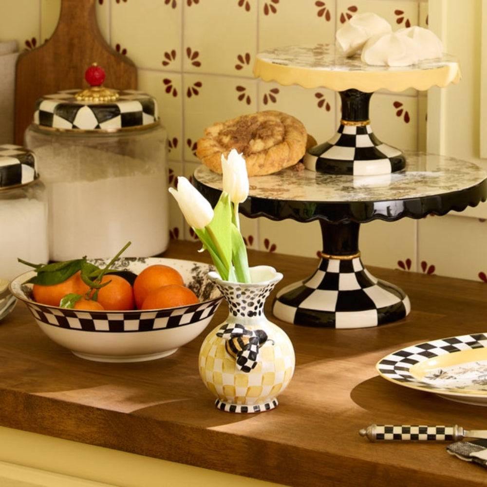 Colorful white, black, green & yellow bee vase with a bee on the front on a petastal with tulips in it on a table with others Mackenzie items in the backround.