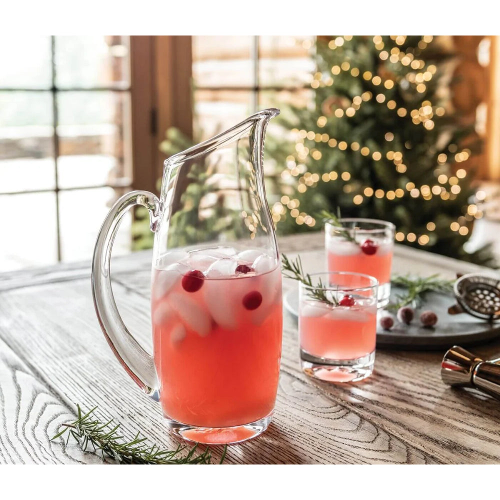 A clear glass pitcher filled with a pink holiday drink, ice, and cranberries, set on a rustic wooden table with two matching glasses and a blurred Christmas tree in the background.