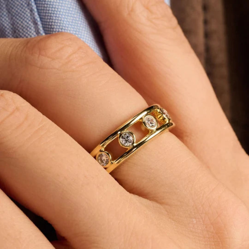 A close-up of a gold ring with bezel-set clear stones worn on a finger. The ring features an open, double-band design with a small gold connector at the back.