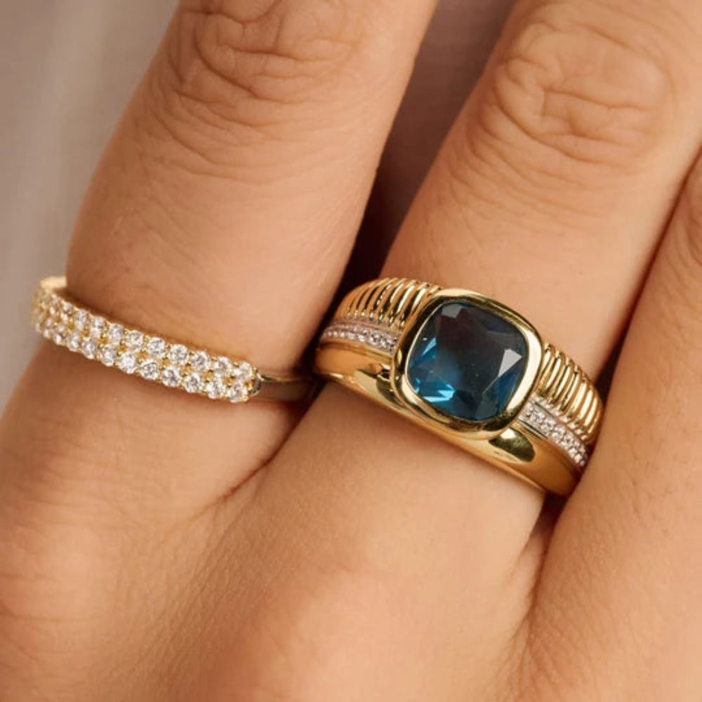 A close-up of a hand wearing two gold rings. The primary ring features a cushion-cut blue gemstone in a ribbed gold setting with small clear stones along the band.