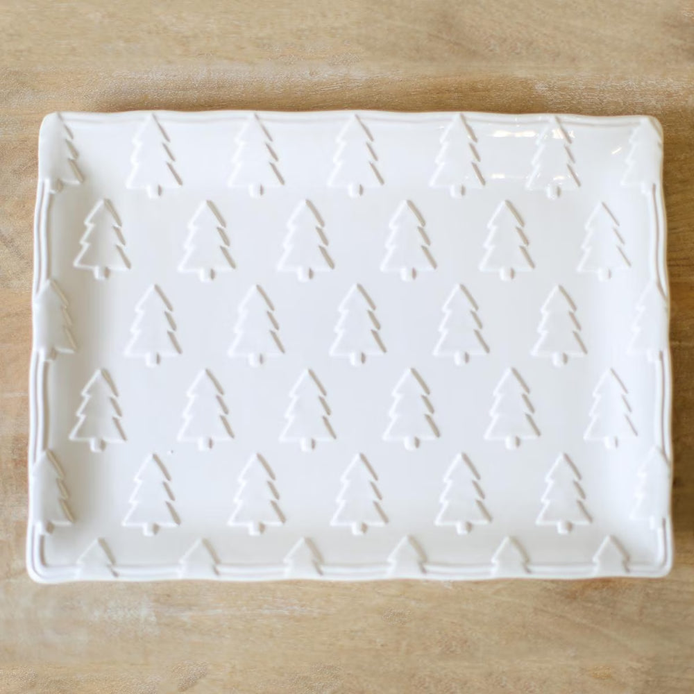 A white rectangular ceramic serving platter with a repeating embossed tree pattern and a wavy rim, shown on a light wood surface.