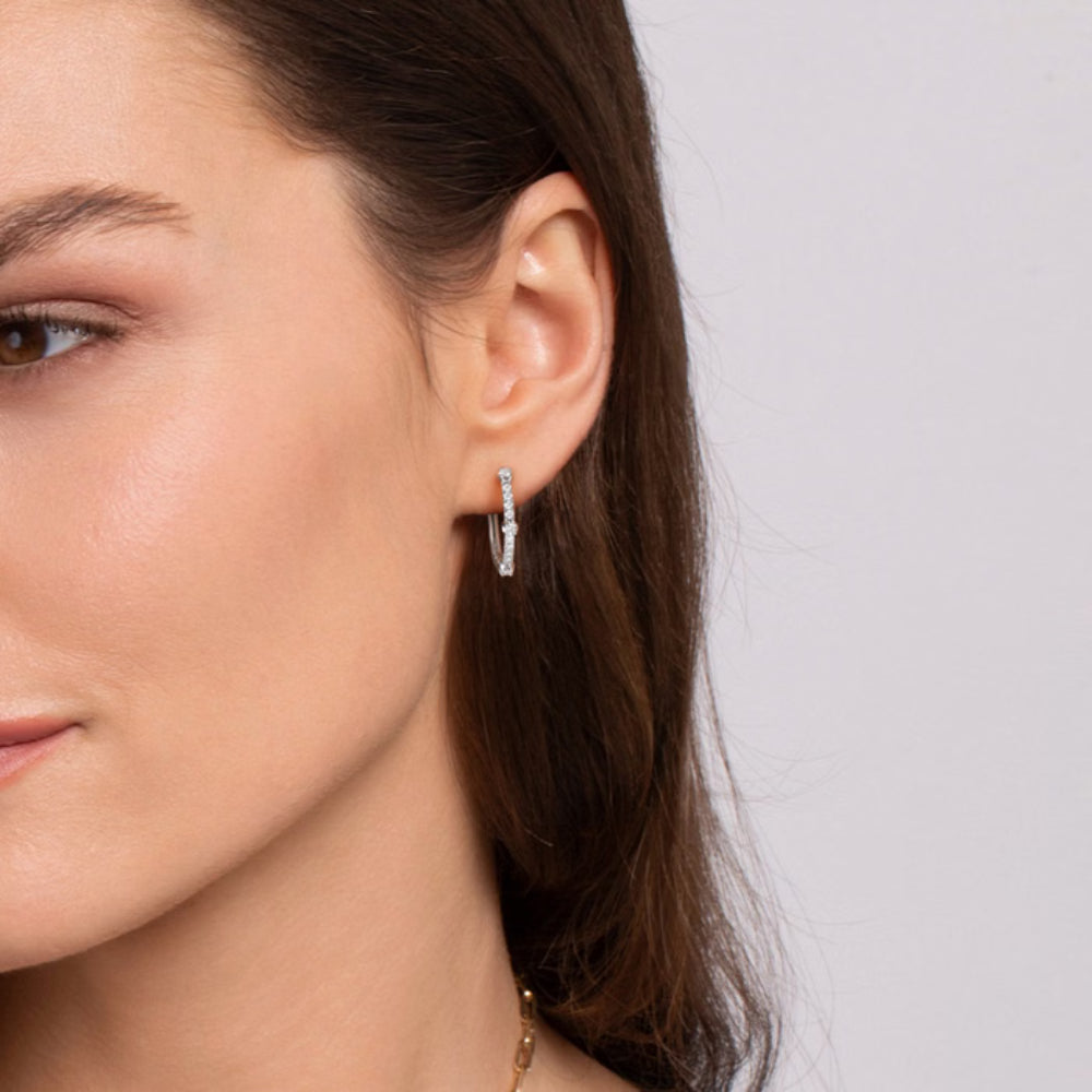A close-up of a woman's ear wearing a delicate gold teardrop-shaped dangle earring with a small clear stone accent, against a light gray background.