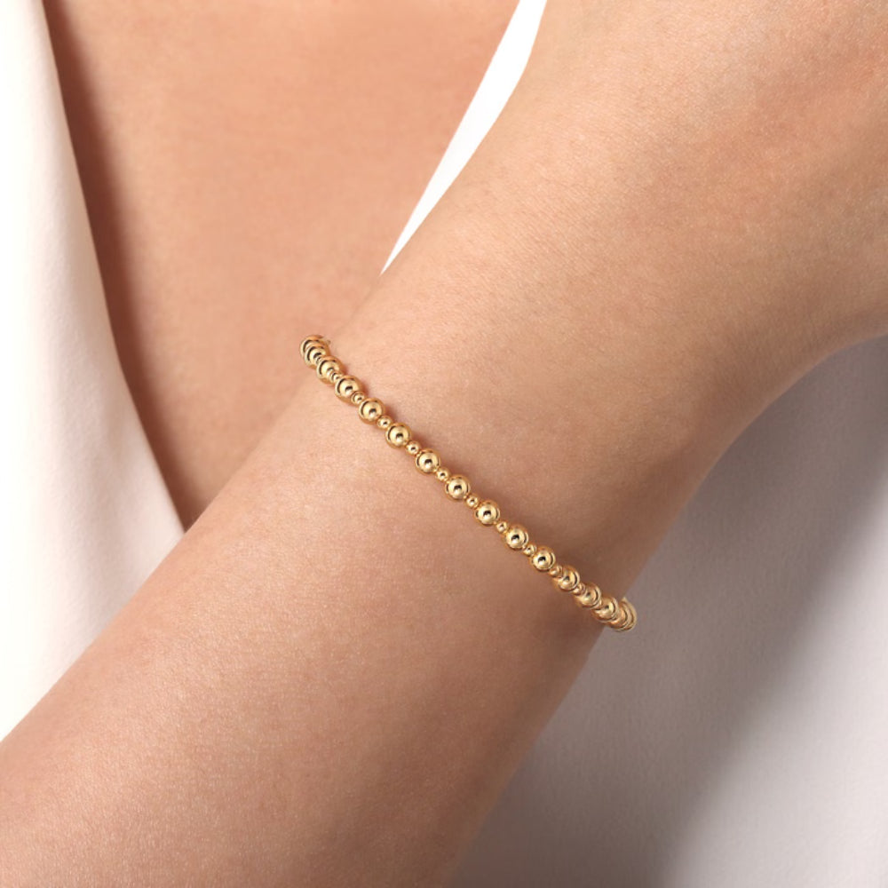A delicate gold beaded bracelet worn on a person's wrist, shown in a close-up shot against a soft, light-colored background.