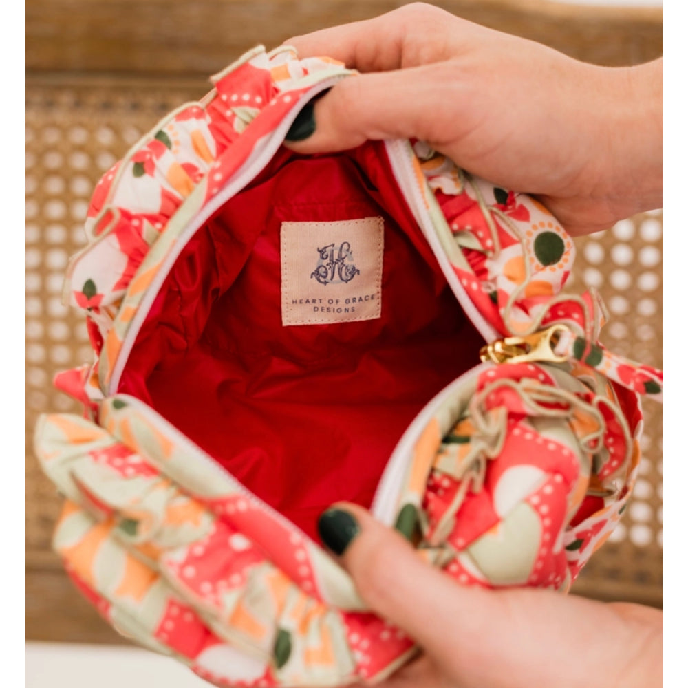A close-up of a fabric clutch bag being held open by hands. The bag has a coral red, orange, and cream floral patterned exterior, a bright red satin lining, and a cream brand label reading 'Heart of Grace Stitchers'.