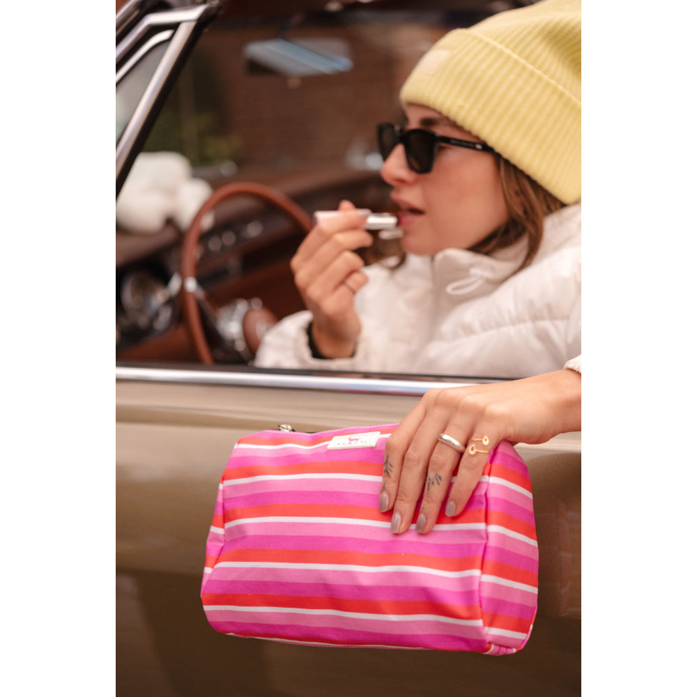 A striped cosmetic pouch in pink, orange, and white horizontal stripes, held by a woman in a yellow beanie and white jacket sitting in a vintage car.