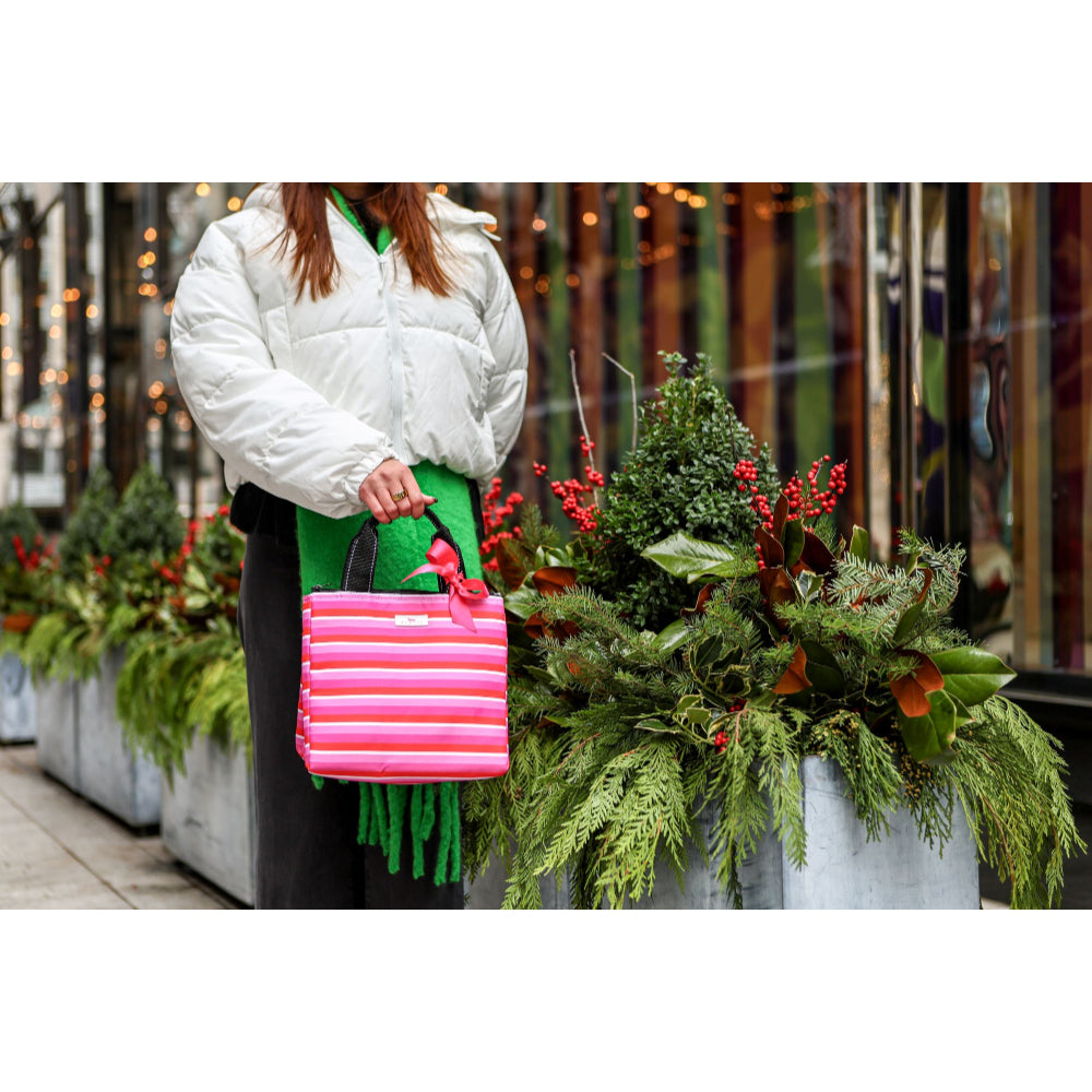 A person in a white puffer jacket and green skirt holds a bright pink striped tote bag with green fringe and a pink bow, standing in front of holiday greenery and window lights.