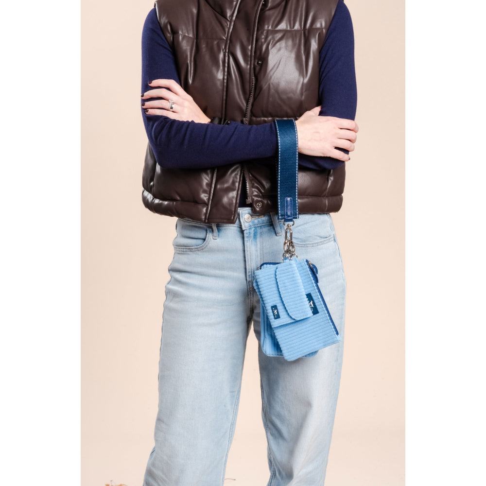 Person wearing a brown puffer vest, blue shirt, light blue jeans, and holding a blue handbag against a beige background.