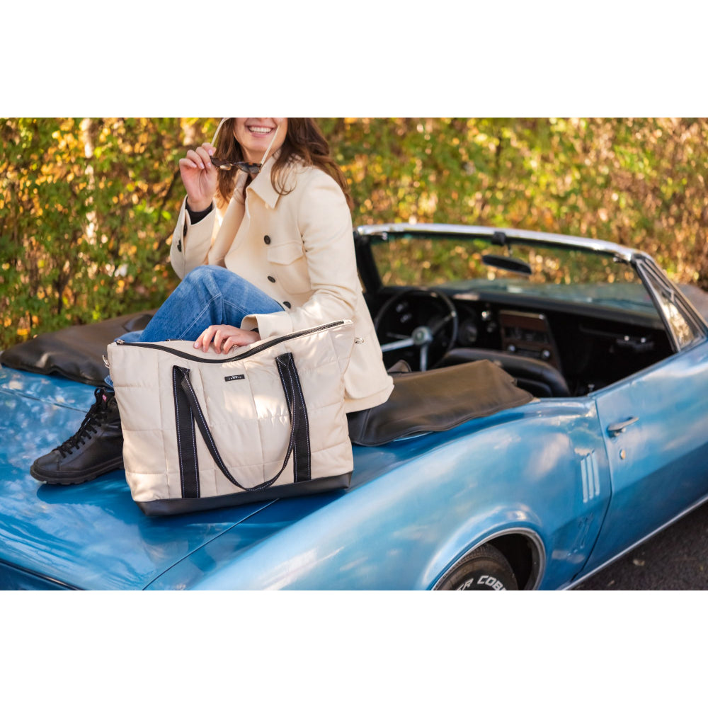 Person sitting on a classic blue car with a white tote bag, surrounded by greenery.