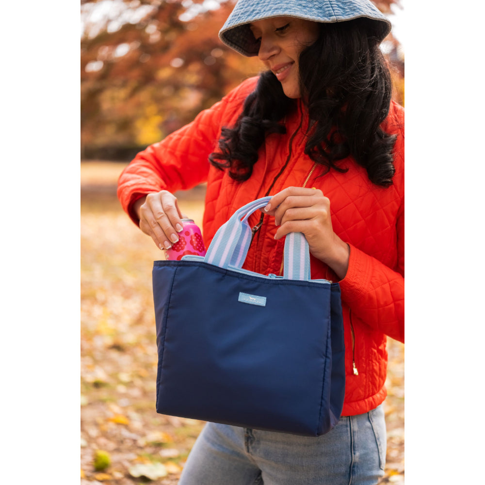 Person in a red jacket holding a blue lunch bag outdoors with autumn leaves in the background