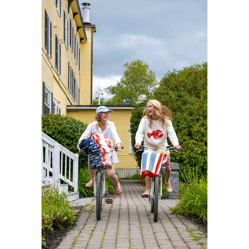 Two women riding bicycles with striped blankets on a paved path in front of a yellow building.