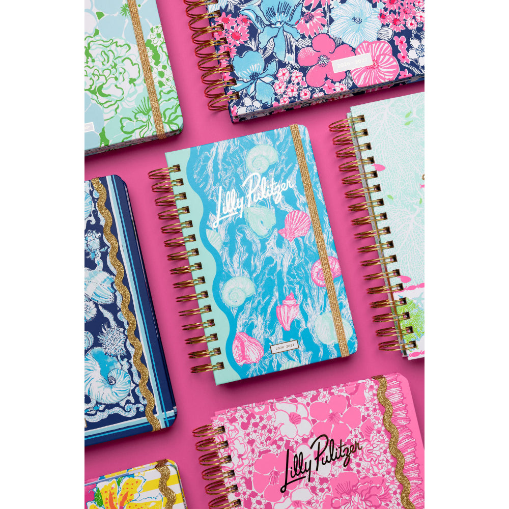 Colorful notebooks with floral and shell patterns on a pink background, featuring the brand 'Lilly Pulitzer'.