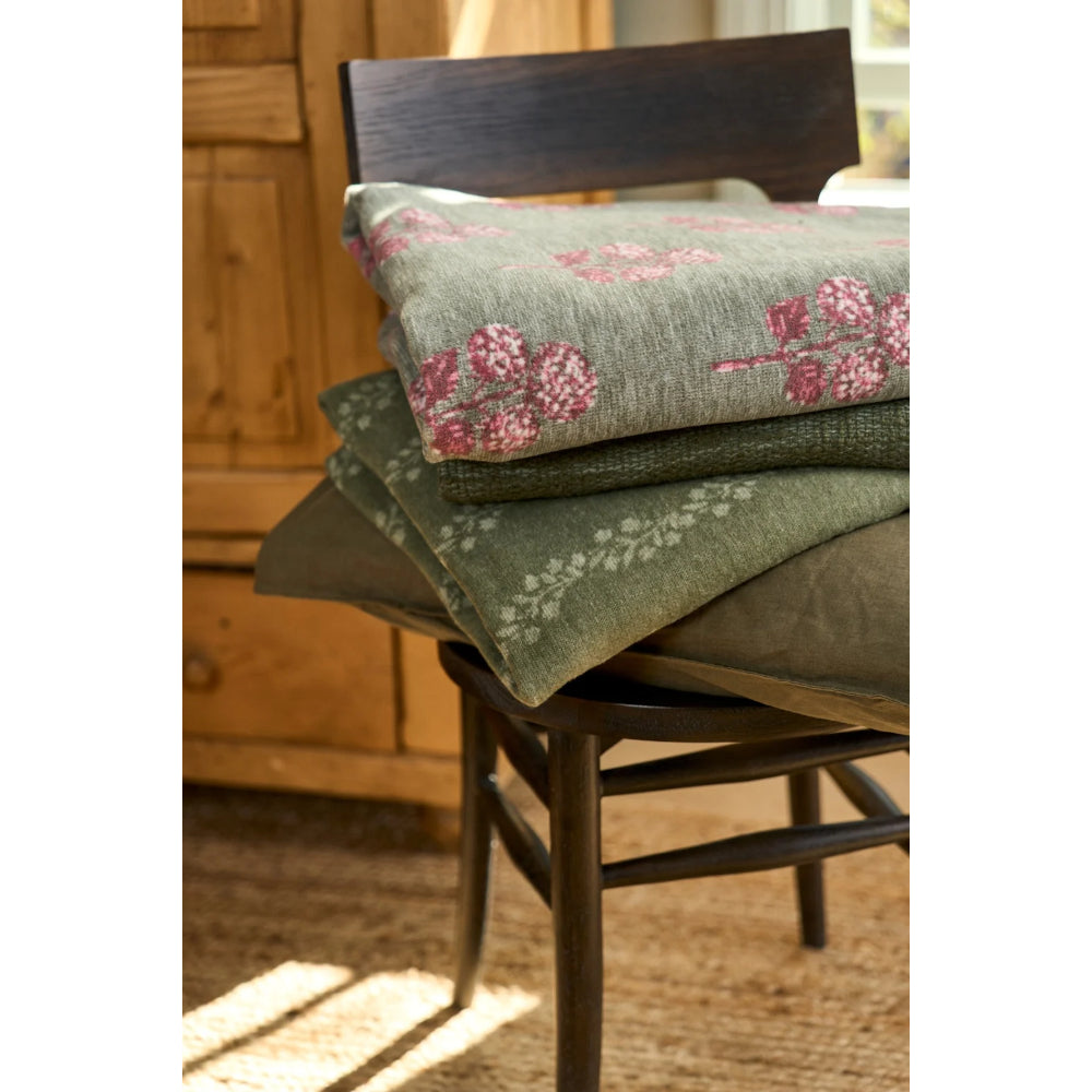 A stack of olive green patterned cushions or throw blankets with pink floral motifs and white floral accents, displayed on a dark wooden chair in a warm indoor setting.