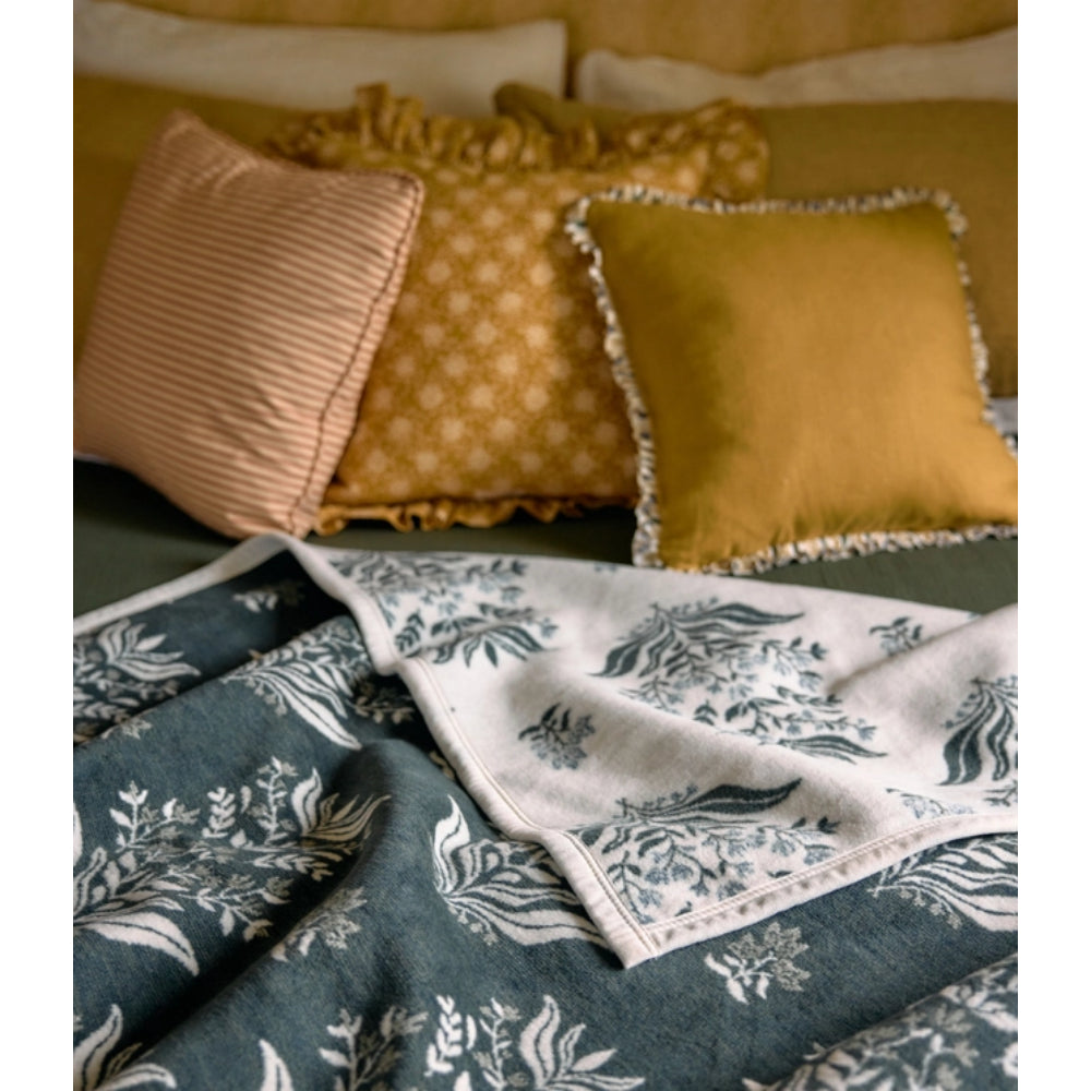 A styled bedding scene with patterned pillows and a folded blanket or throw in front of a bed. The visible textiles include a dark teal blanket with a white botanical print, a white cloth with a blue floral border, and mustard and blush pillows.