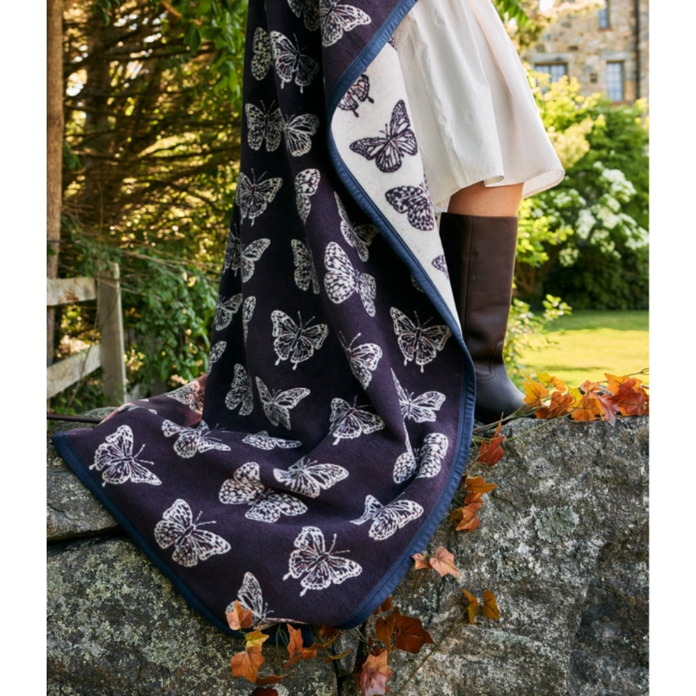 A dark navy blanket or throw with a white butterfly pattern draped over a person's lap and leg, with a white blouse and dark brown knee-high boots visible, set outdoors on a stone wall with autumn leaves and a garden background.