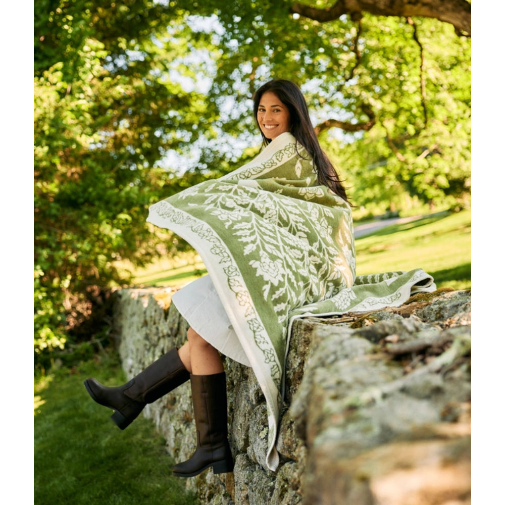A woman sitting on a stone wall outdoors, wrapped in a large sage green and white patterned blanket. She is wearing dark brown knee-high boots, with a grassy park and trees in the background.