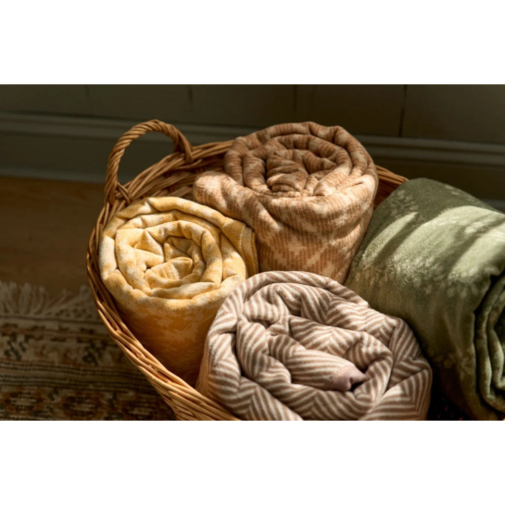 Three rolled plush blankets in beige, brown, and sage green tones arranged inside a wicker basket on a patterned rug.