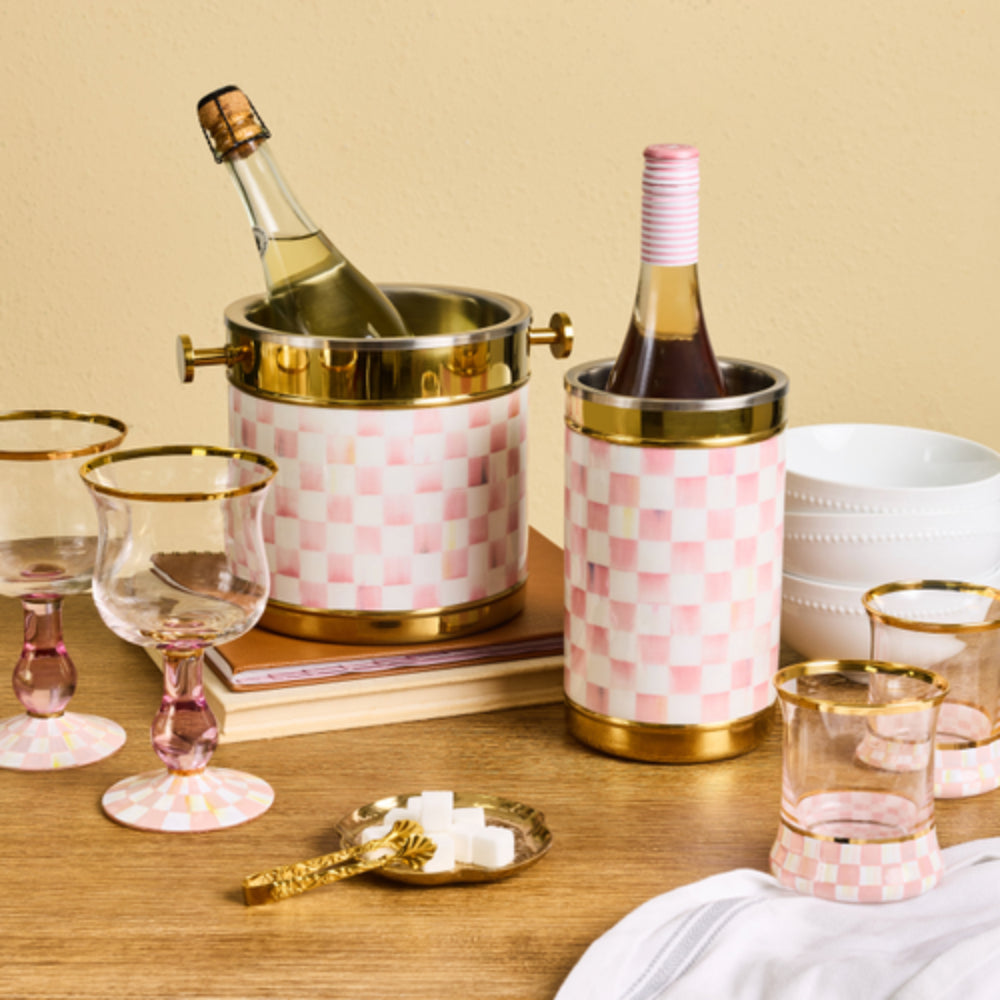 cylindrical metal bucket-style container with a pink and white checkerboard patterned body, gold-toned top and bottom bands, and small side handles on a wood table with glasses and wine bottles in it with other servware.