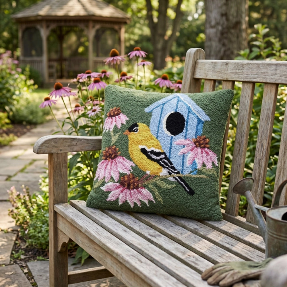 Decorative pillow with bird and birdhouse design on a wooden bench in a garden.