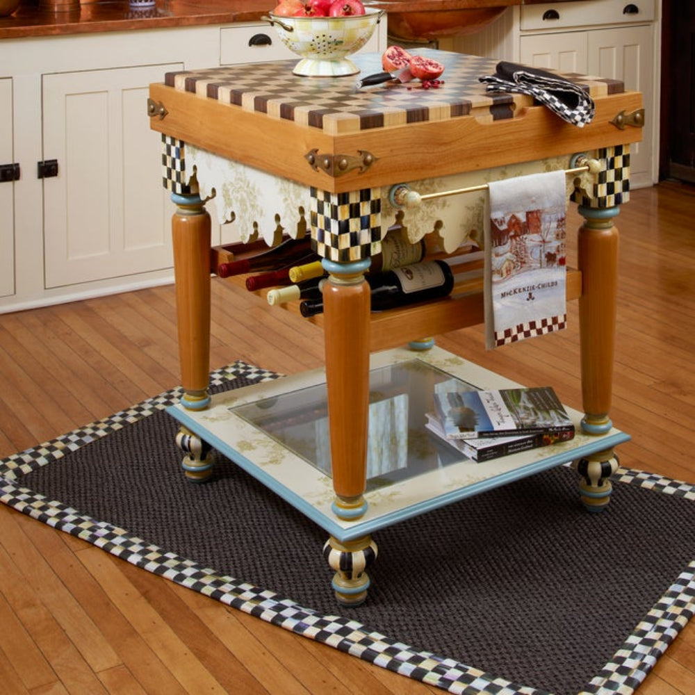 Life style photo of MacKenzie-Childs courtly check black sisal rug with black checkered border on the floor in a kitchen with a wood floor with a wood table with black checks on it.