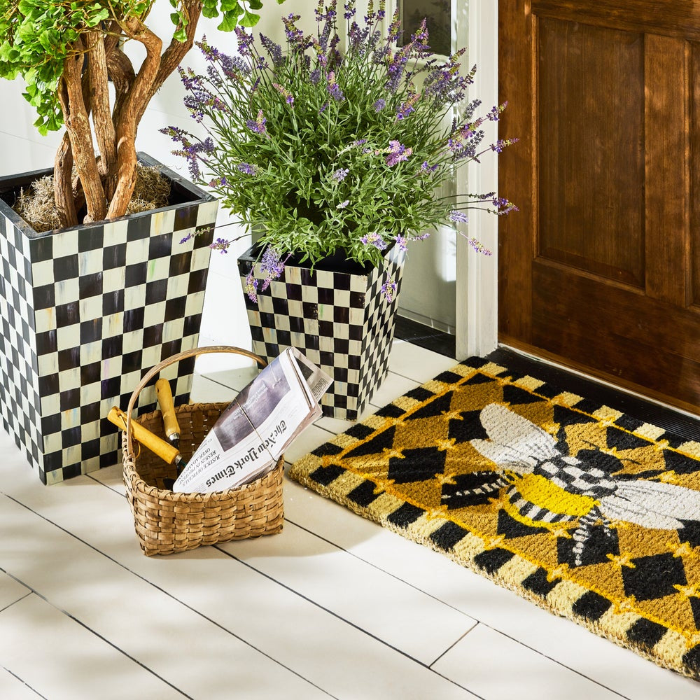 Decorative entrance with checkered planters, a basket, and a doormat with a bee design.