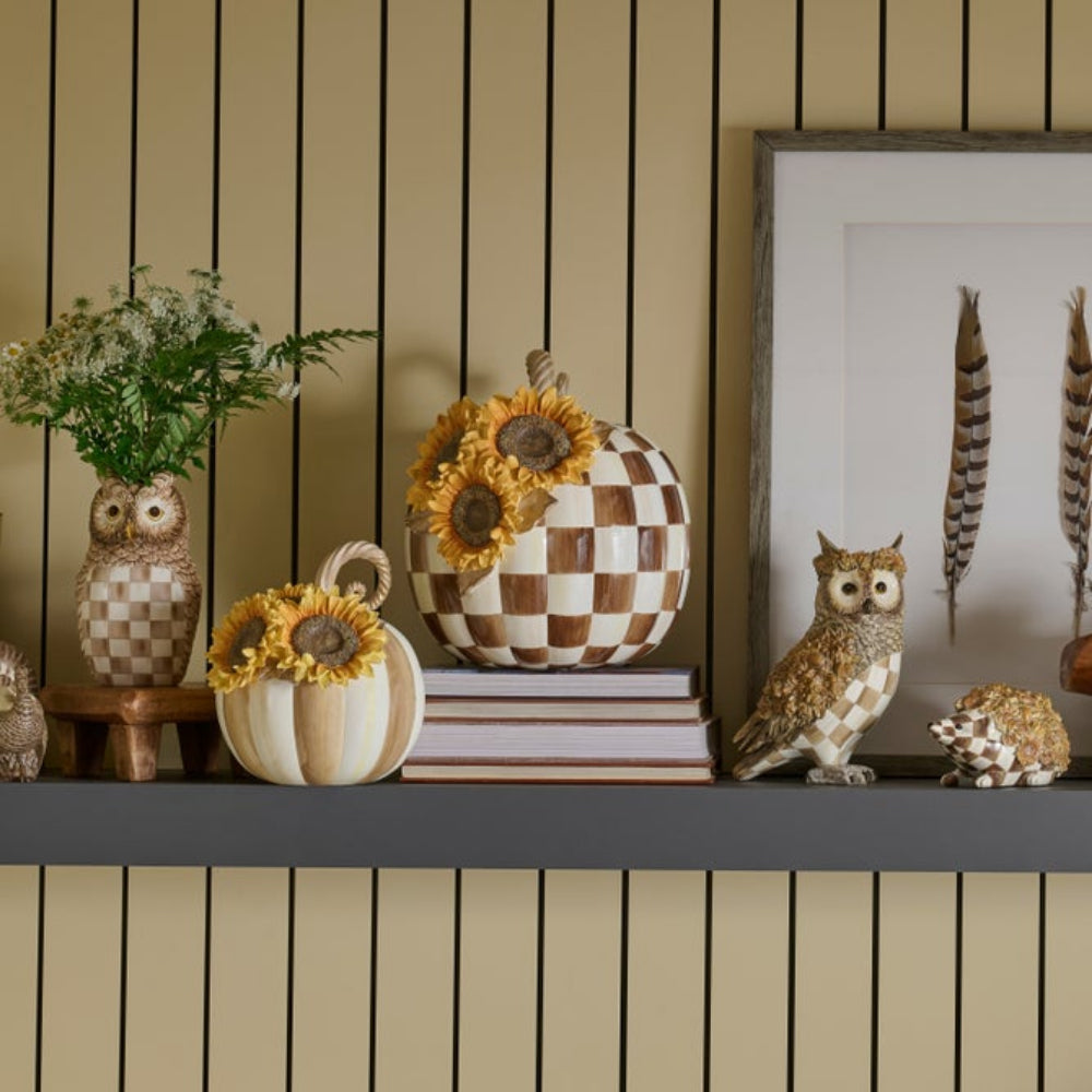 Autumn-themed home decor collection displayed on a dark gray shelf against a beige wood-paneled wall, featuring checkered pumpkins, sunflower vases, owl figurines, a hedgehog ornament, and a framed feather print.