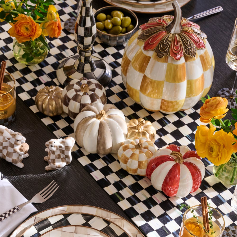 A styled autumn tabletop display with decorative ceramic pumpkins in white, gold, red, and brown checkerboard patterns, arranged on a black-and-white checkered runner with flowers, plates, and glasses.