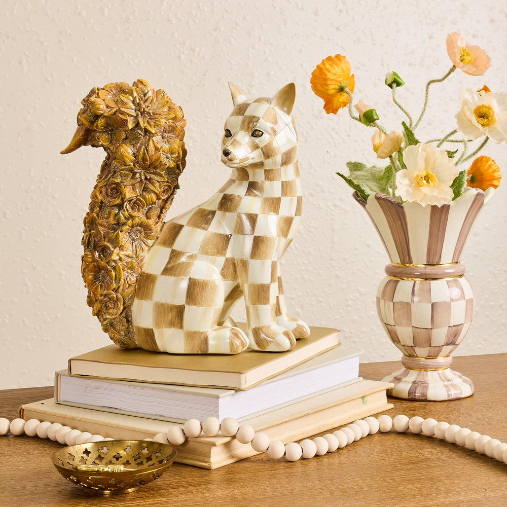 A decorative ceramic fox figurine with a beige and brown checkerboard pattern sits on top of stacked books, next to a matching patterned vase holding orange and cream flowers. A gold-toned bowl and a beaded garland are also visible on a wooden table against a textured beige wall.