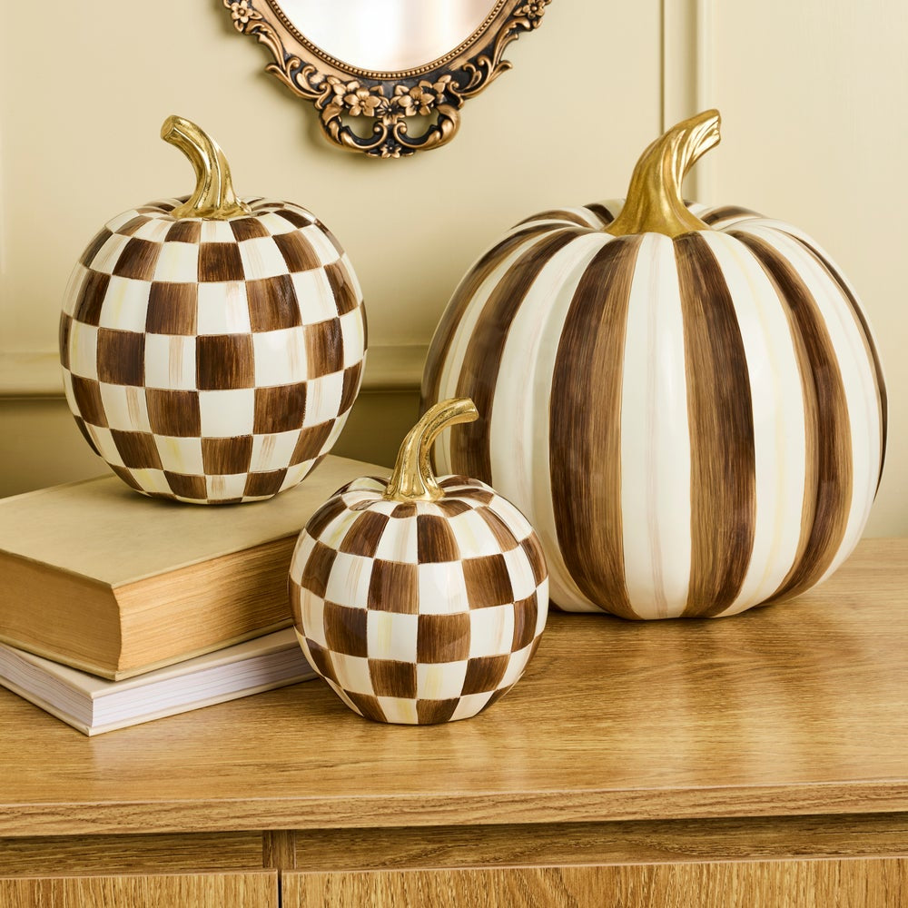 Three decorative pumpkins displayed on a wooden table against a beige wall. One large pumpkin has vertical brown and cream stripes, one smaller striped pumpkin is partially visible behind a stack of books, and one small pumpkin has a brown and cream checkerboard pattern.