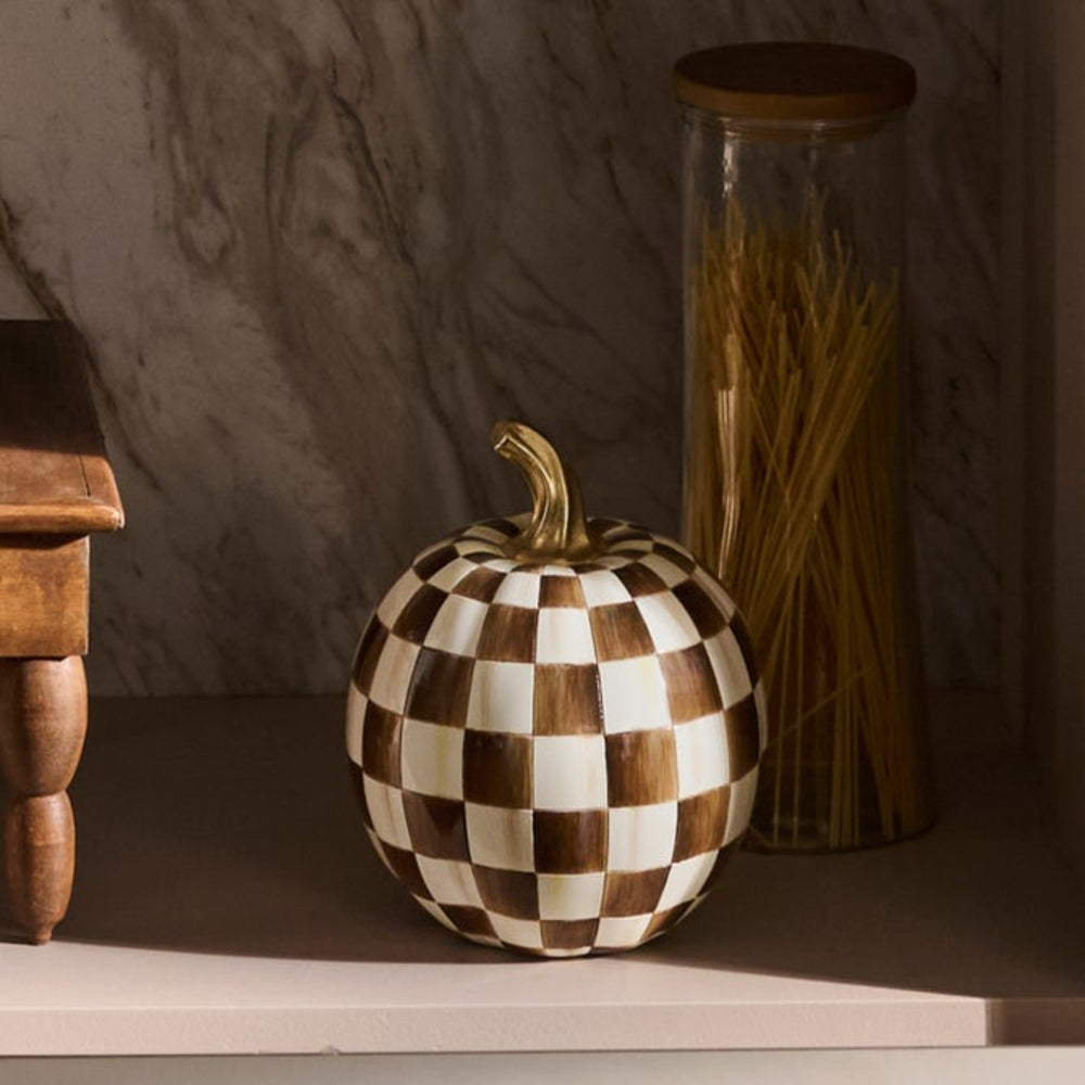 A decorative pumpkin-shaped container with a brown and cream checkerboard pattern and a gold-toned stem, displayed on a shelf next to a glass jar of spaghetti.