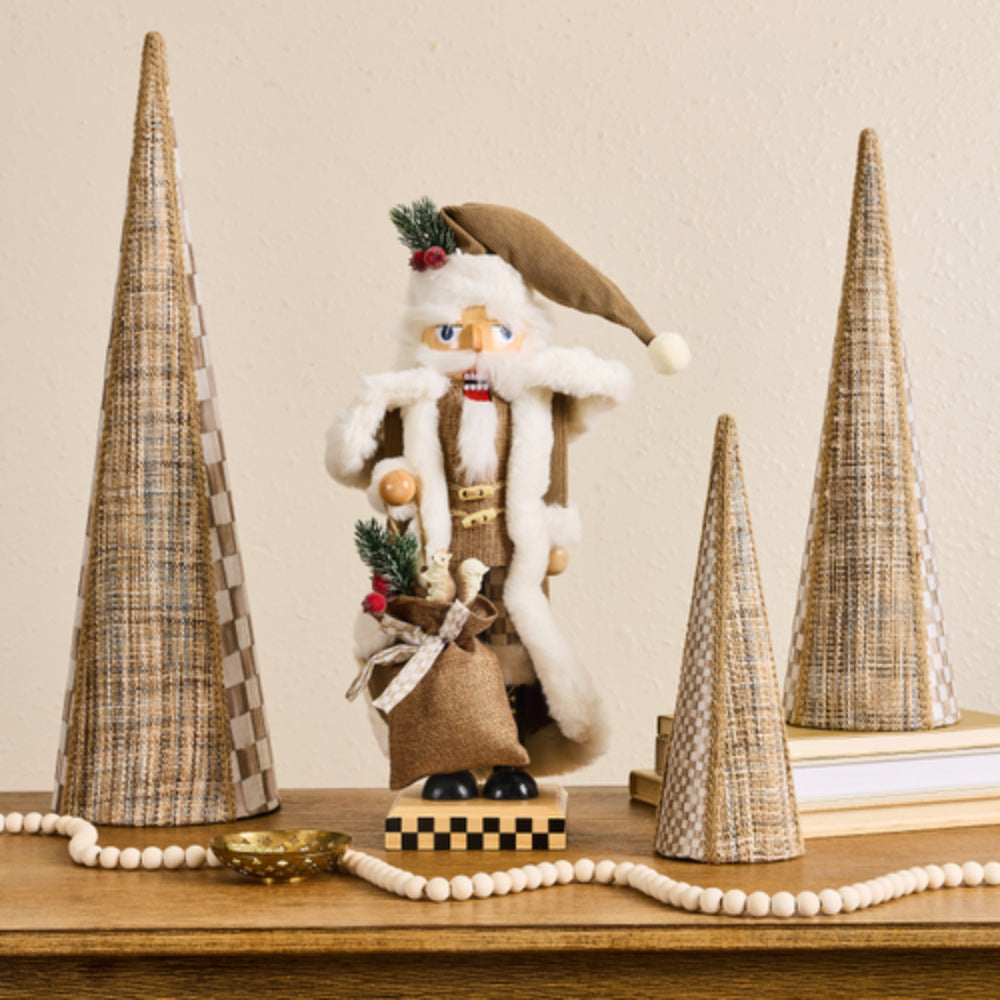 A festive Christmas nutcracker figurine standing on a wooden table, flanked by three tall woven cone decorations, a beaded garland, and a small gold bowl against a warm beige wall.