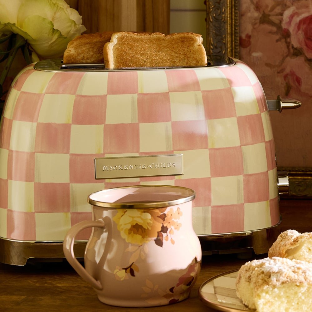 A retro-style pink and cream checkered toaster with two slices of toast, shown in a warm kitchen setting with a floral mug in the foreground and baked goods on a plate.