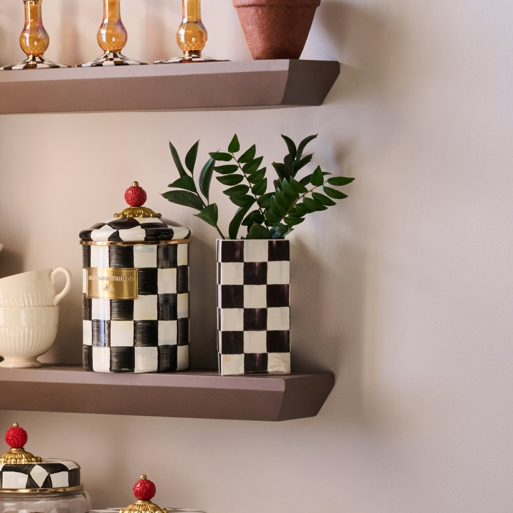 A black and white checkered ceramic canister with a gold band and red top, displayed on a brown floating shelf next to a matching checkered rectangular planter holding green leaves, against a warm beige wall.