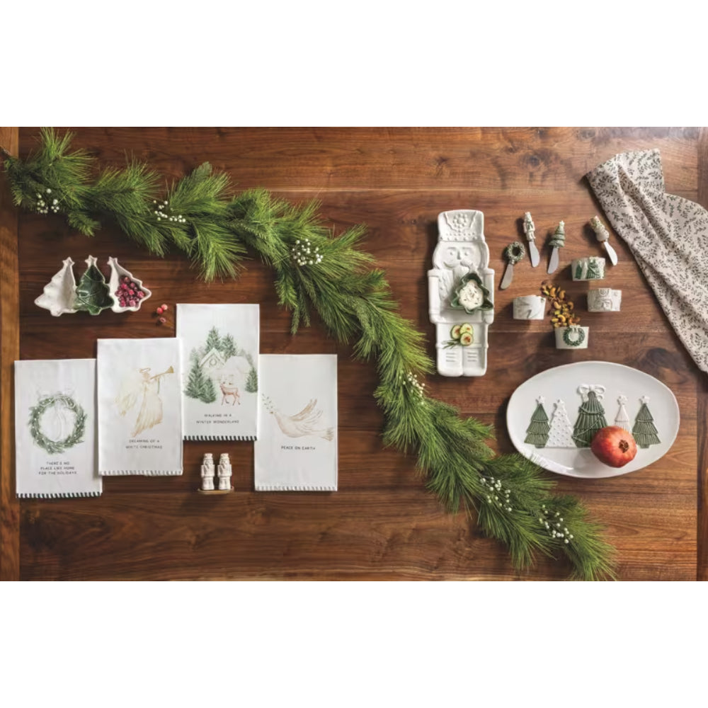 A styled flat-lay of Christmas-themed table linens and serving pieces arranged on a wooden table, including printed napkins or tea towels with holiday illustrations, a white divided serving tray, small dishware pieces, and a patterned cloth. A green garland and a red apple are placed among the items.