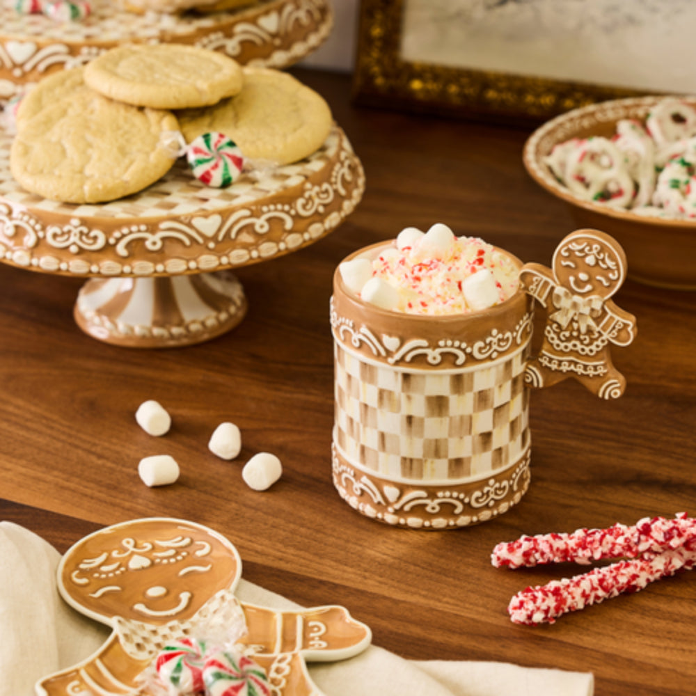 A gingerbread-themed ceramic mug with a checkered pattern and gingerbread man handle, filled with marshmallows and topped with candy canes, set on a wooden table with cookies and candy props.