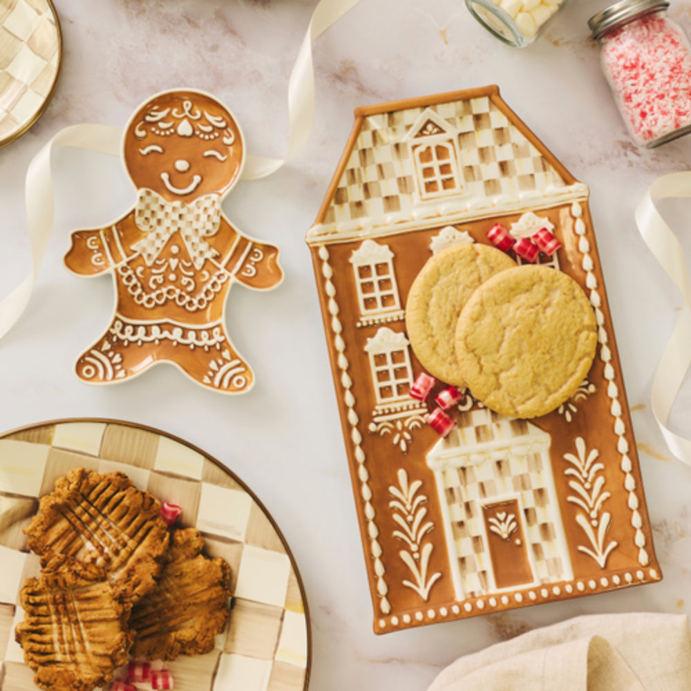 Gingerbread house shaped cookie cutter and tray with decorated gingerbread cookies, arranged on a white marble surface with ribbon and jars in the background.
