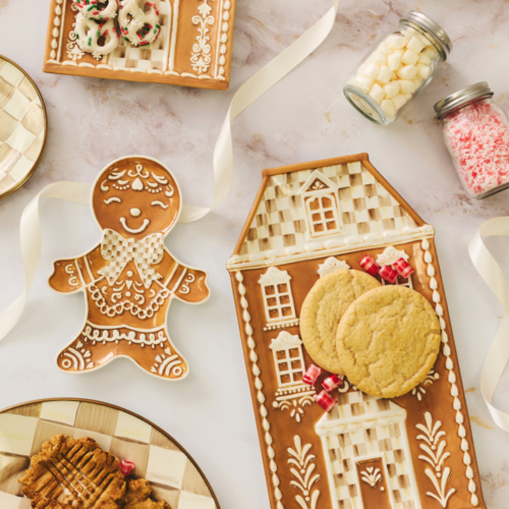 A styled flat-lay of gingerbread-themed cookie dishes on a light marble surface, including a gingerbread man shape plate and a house-shaped plate holding two cookies, with candy jars and ribbons around them.