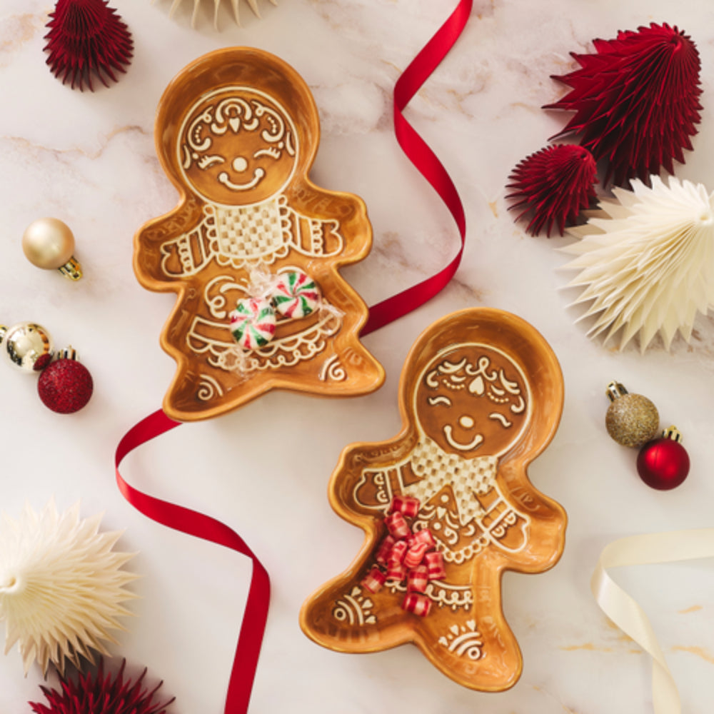 Two gingerbread man-shaped ceramic serving dishes decorated with icing-like patterns, arranged on a light marble surface with red ribbons and Christmas ornaments.
